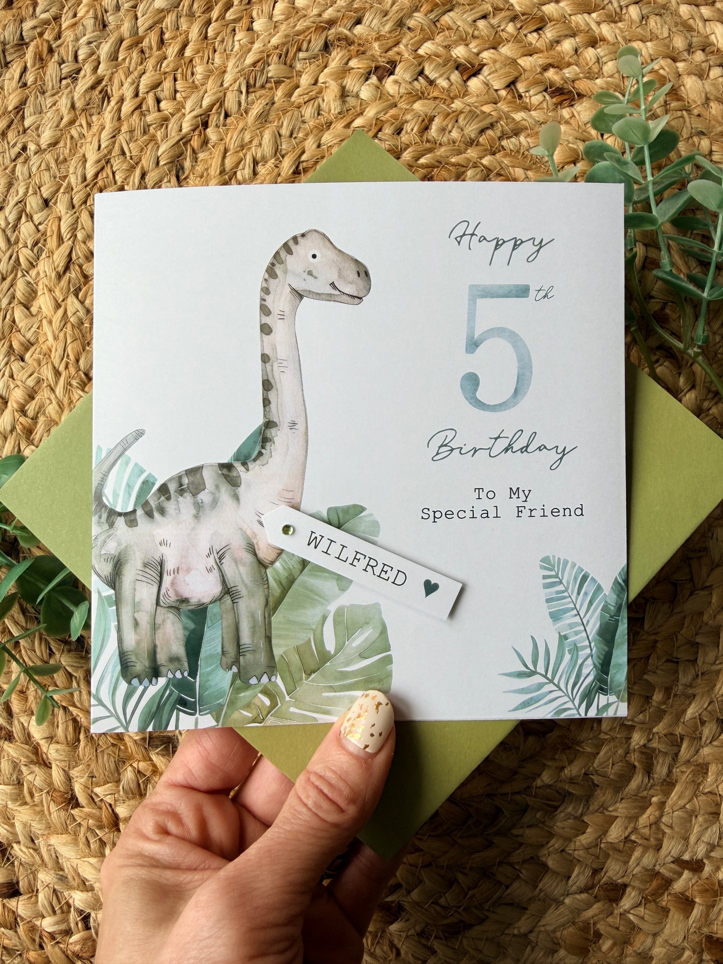 Hand holding a birthday card with a dinosaur illustration on a woven surface