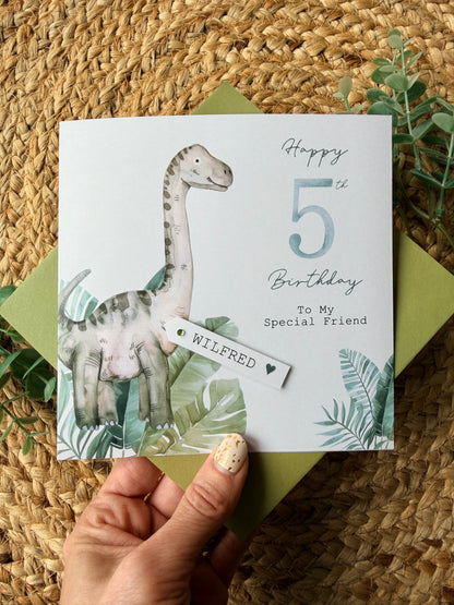 Hand holding a birthday card with a dinosaur illustration on a woven surface