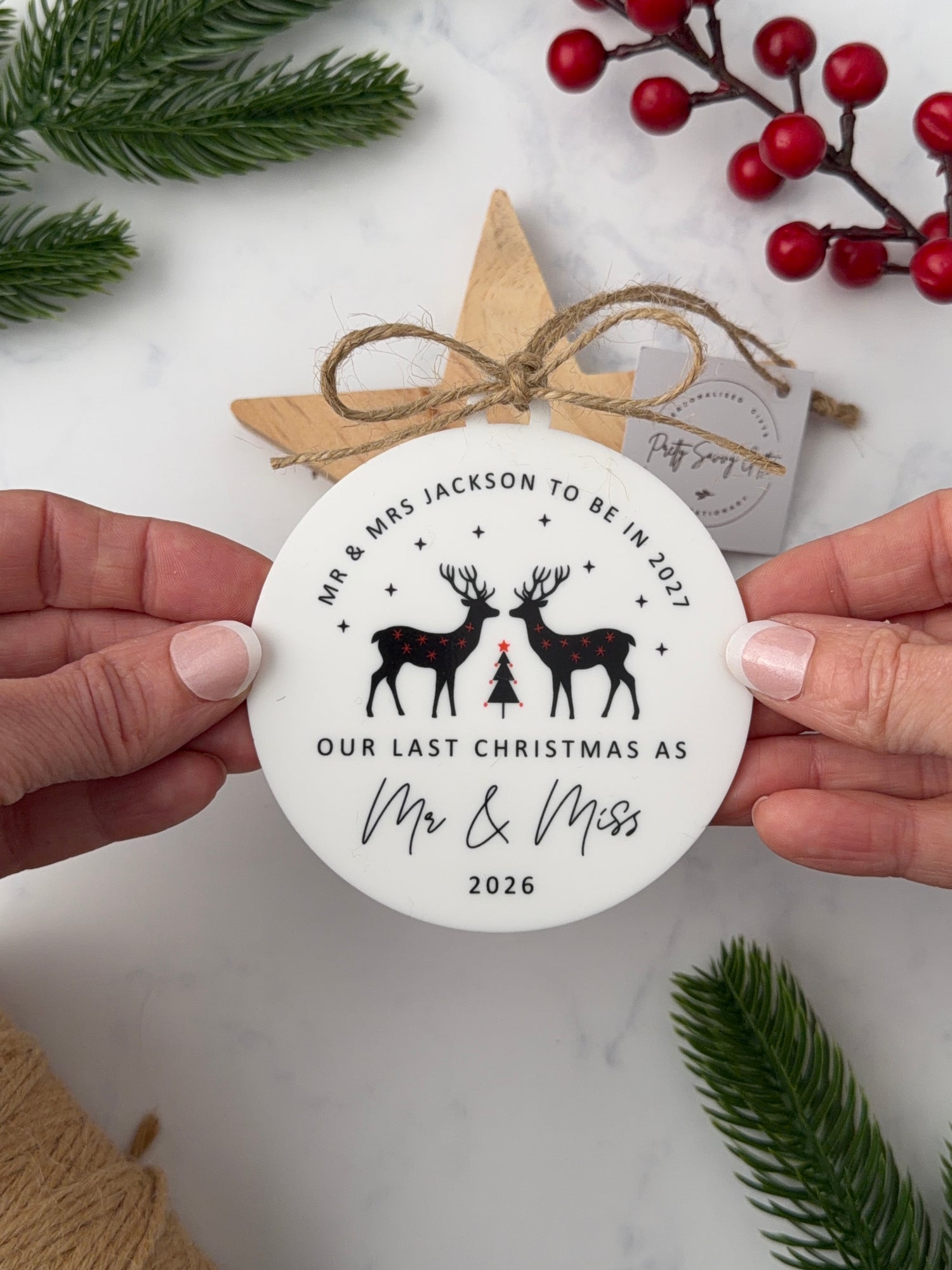 A person holds a round white personalized Christmas ornament or favor tag with black reindeer silhouettes and printed text, surrounded by holiday props including pine branches, red berries, a wooden star, and a gift tag on a light marble surface.