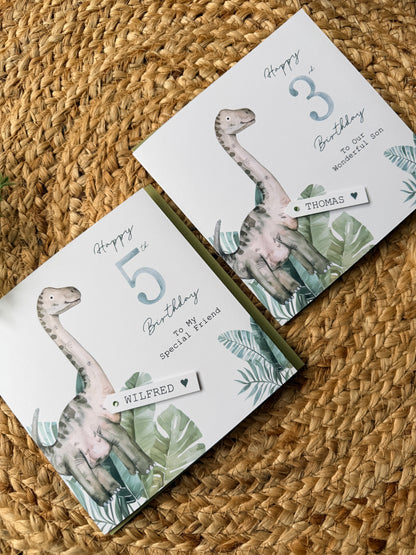 Two birthday cards with dinosaur illustrations on a woven surface