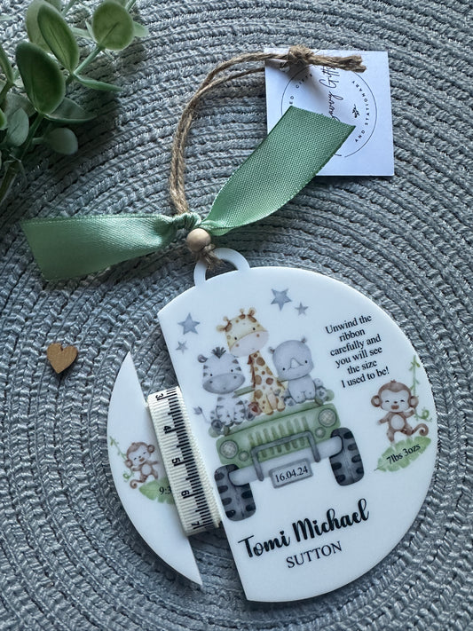 A personalized baby keepsake plaque featuring an illustration of a green jeep, baby safari animals and text with the baby's name, date of birth, weight and height if know. The white plaque is decorated with a sage green ribbon and a small wooden bead, and is shown with its custom printed gift tag on a textured grey woven background.