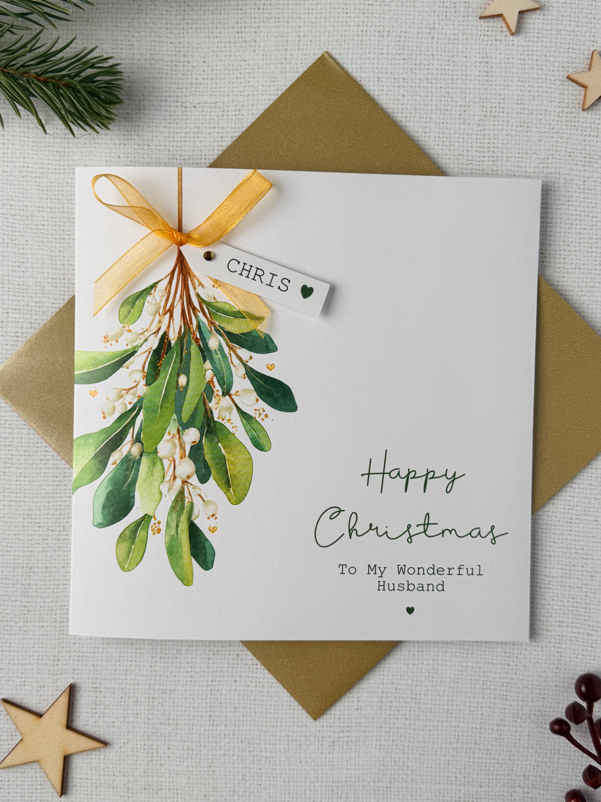 A personalised Christmas greeting card with a watercolor mistletoe illustration, a gold ribbon, and a small hanging tag with name and placed on top of a gold envelope on a light fabric background with wooden star decorations.