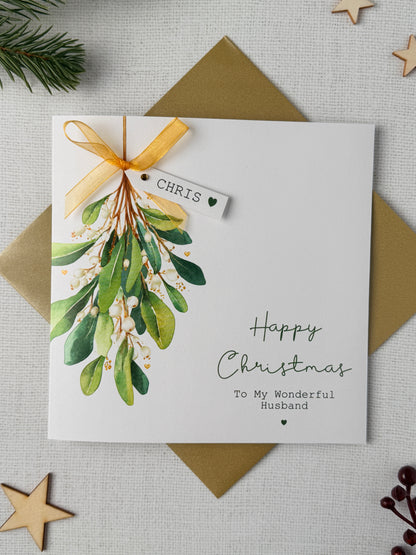 A personalised Christmas greeting card with a watercolor mistletoe illustration, a gold ribbon, and a small hanging tag with name and placed on top of a gold envelope on a light fabric background with wooden star decorations.