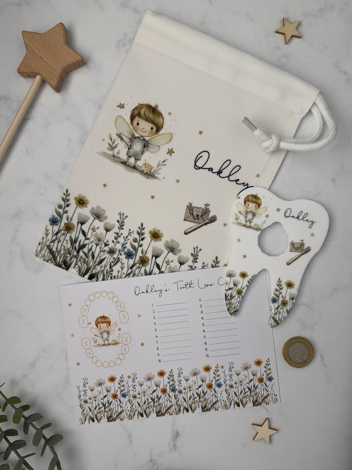 A white fabric tooth fairy bag with a matching printed tooth chart and acrylic shaped coin tooth holder, styled on a light marble surface with wooden star decorations and greenery.
