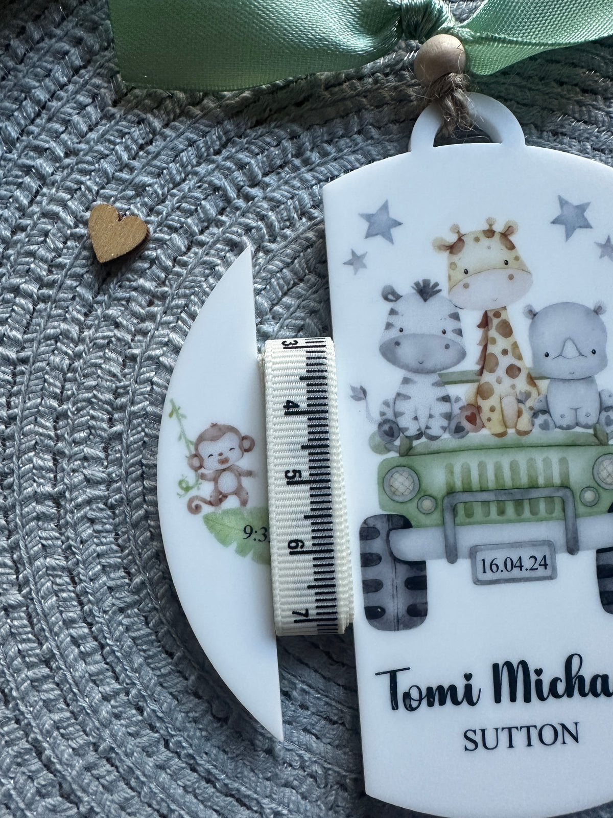 A close-up of a personalized keepsake tag or ornament with cute animal illustrations and the text of the baby's name, shown with a small fabric measuring tape and a ribbon on a grey woven surface.