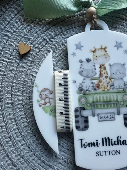 A close-up of a personalized keepsake tag or ornament with cute animal illustrations and the text of the baby's name, shown with a small fabric measuring tape and a ribbon on a grey woven surface.