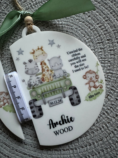 A personalized acrylic baby keepsake plaque shaped like a safari vehicle, featuring illustrated animals, a green jeep, stars, and the name beside it. A green ribbon is tied to the side, and a measuring tape is placed along the left hand edge.