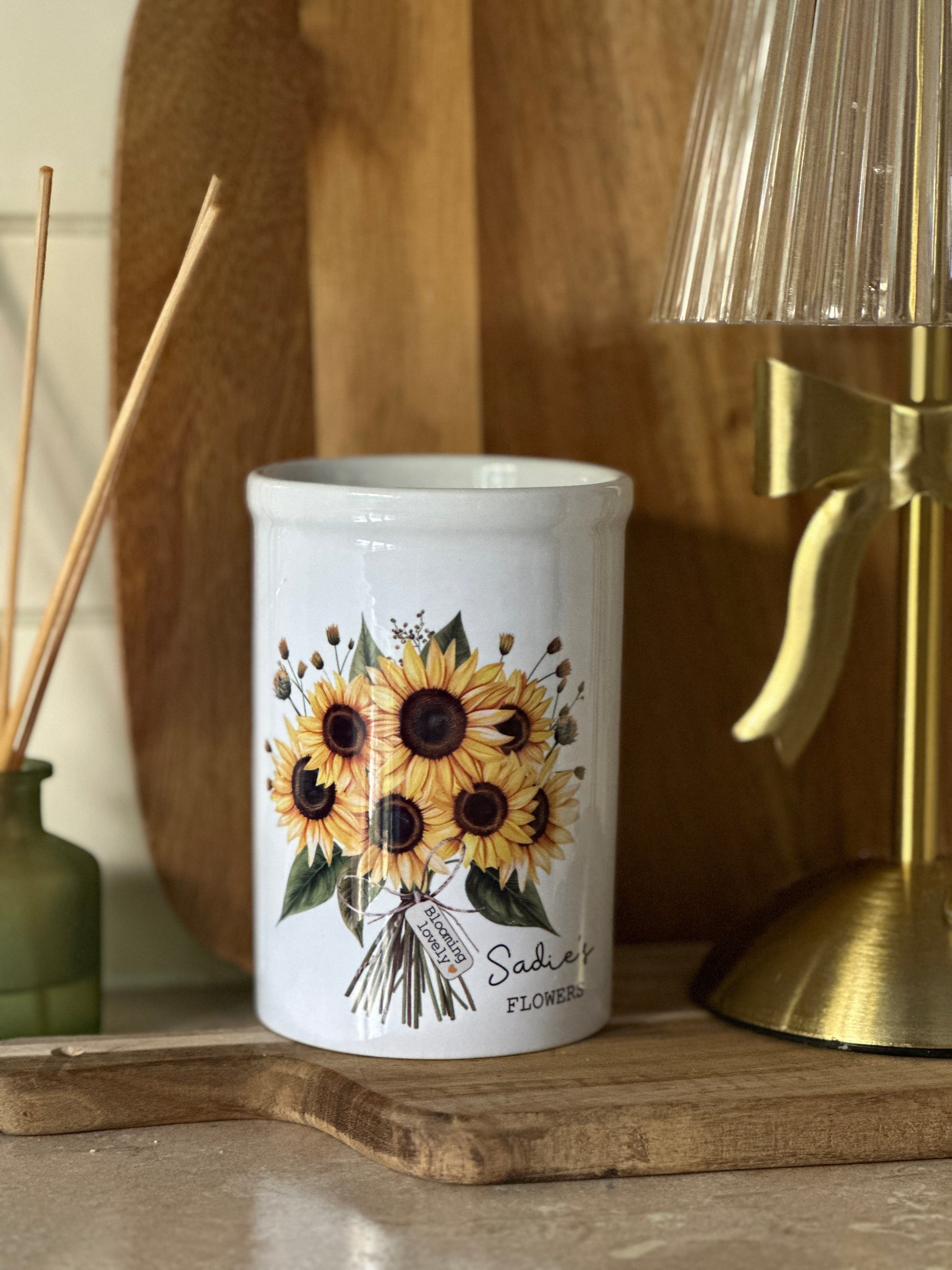White personalised vase with sunflower design on a wooden surface with a gold lamp and reed diffuser in the background.