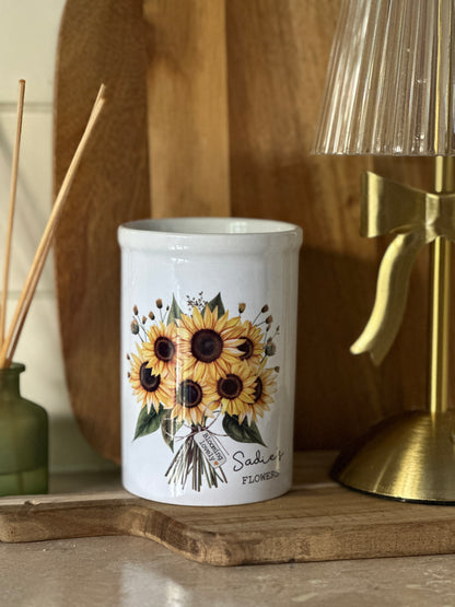 White personalised vase with sunflower design on a wooden surface with a gold lamp and reed diffuser in the background.